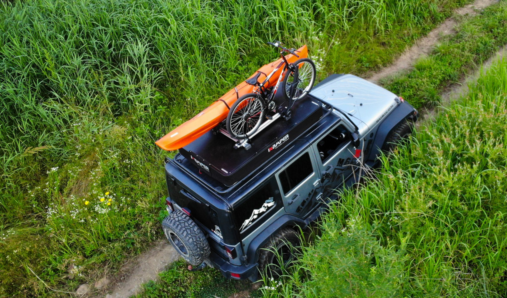 Een terreinwagen staat op een veldweg. Op het dak zijn een harde schaal daktent voor 3 personen, een fiets en een kano bevestigd.