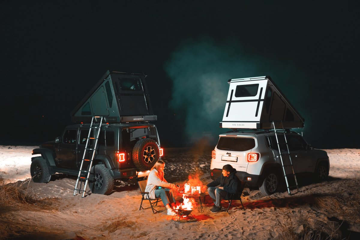 Twee personen zitten op campingstoelen bij het kampvuur. Op de achtergrond staan hun auto's met Dark Barracks Hardside daktenten