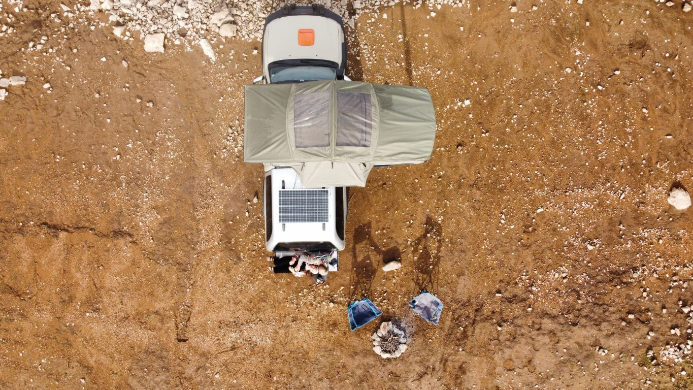 Adventure daktent van Campwerk vanuit vogelperspectief. Een stel zit achter de auto, daarnaast staan kampeerstoelen en een vuurplaats 