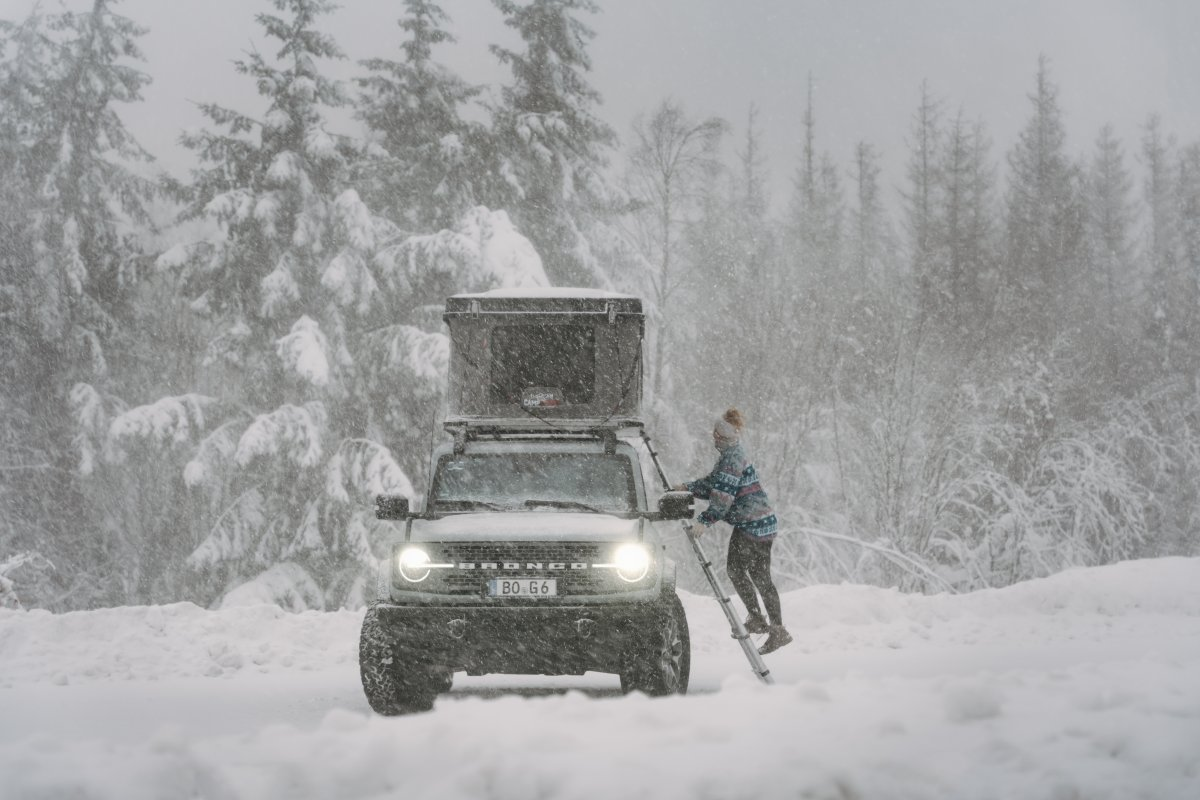 Vrouw klimt via de ladder bij sneeuwstorm in haar hardshell daktent van CAMPWERK